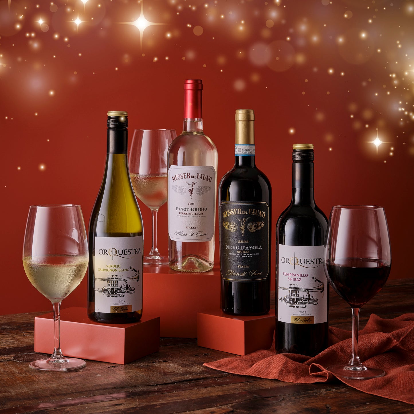 Four wine bottles with labels and two glasses on a wooden surface against a warm, blurred background.