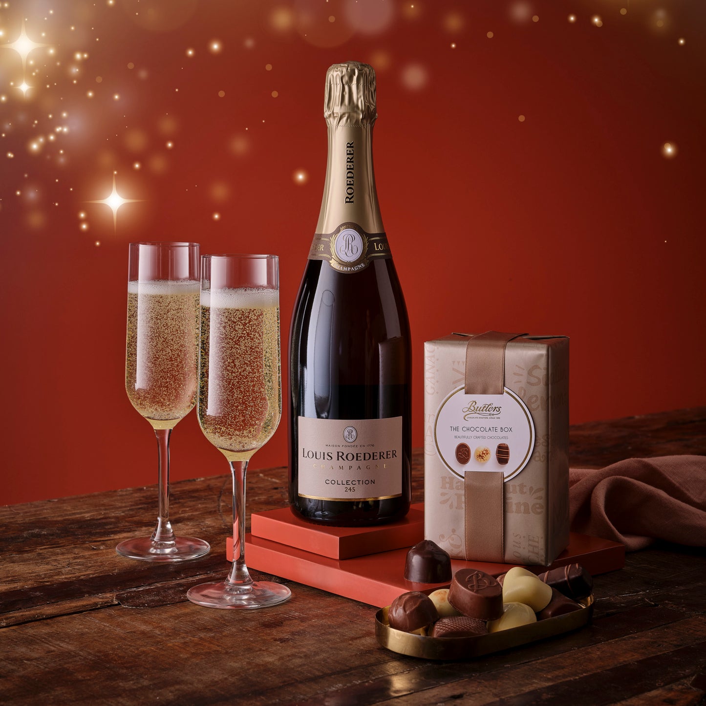 Bottle of Louis Roederer champagne with two glasses and a box of chocolates on a wooden surface.