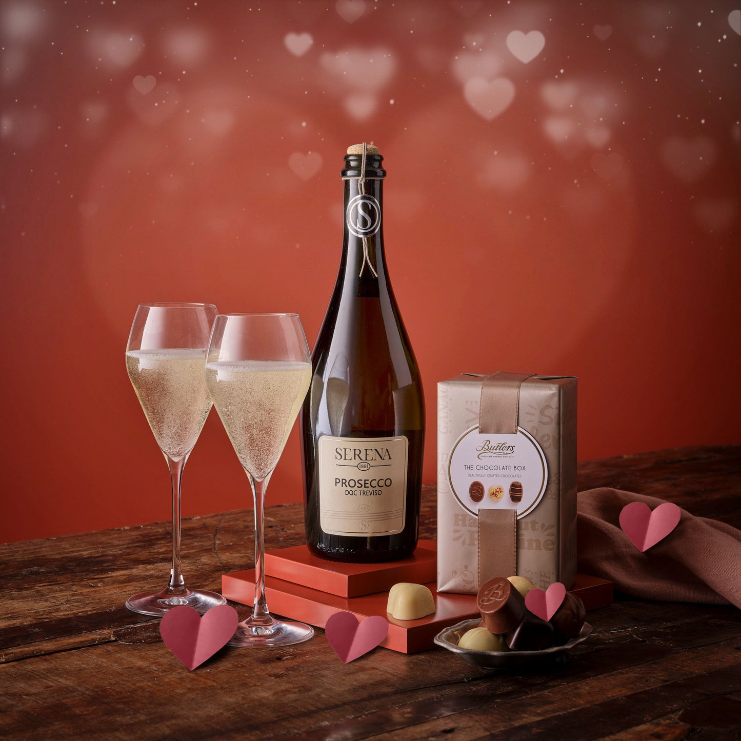 Prosecco and chocolates hamper with two champagne glasses, a Serena Prosecco bottle, a chocolate box, and pink hearts on a rustic wooden table.