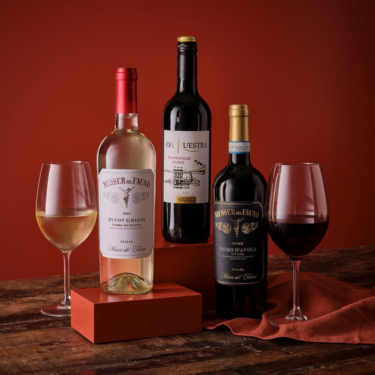 Three mixed wine bottles Pinot Grigio Orquestra Tempranillo Shiraz Nero d'Avola with two glasses on a warm red backdrop.