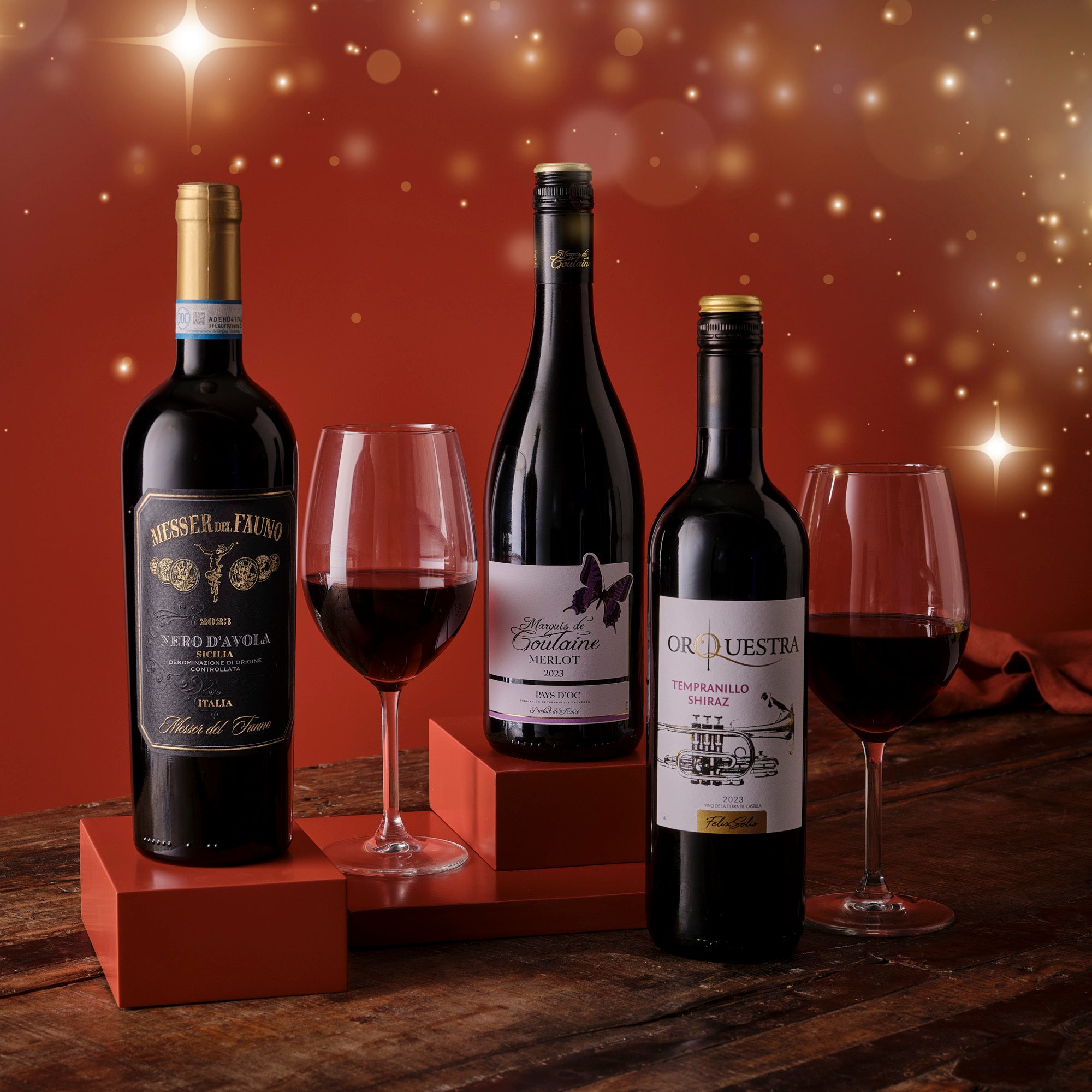 Three bottles of red wine with glasses on a wooden surface against a warm, blurred background.