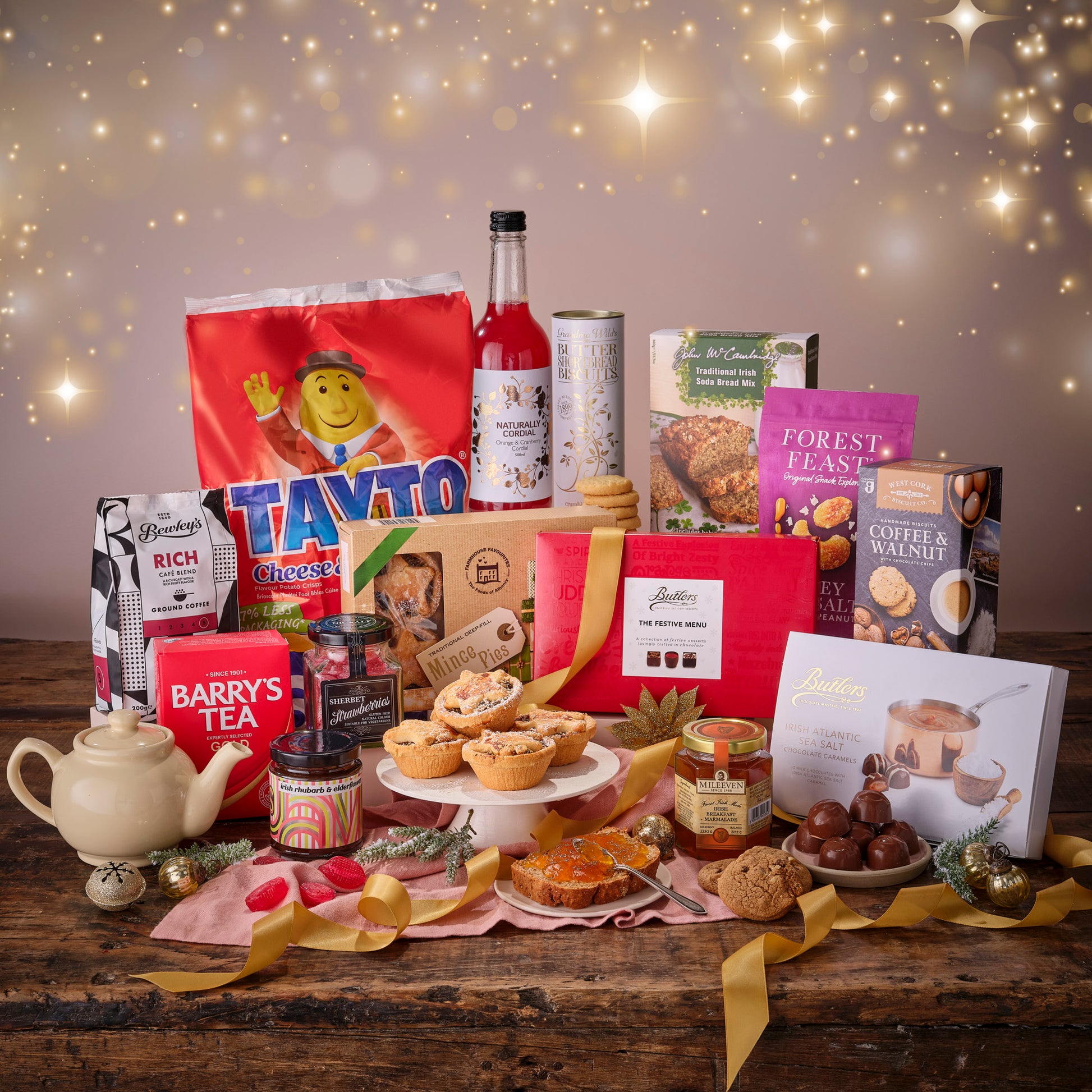 Assorted food products including tarts, jars, and a teapot on a wooden table with a festive background.