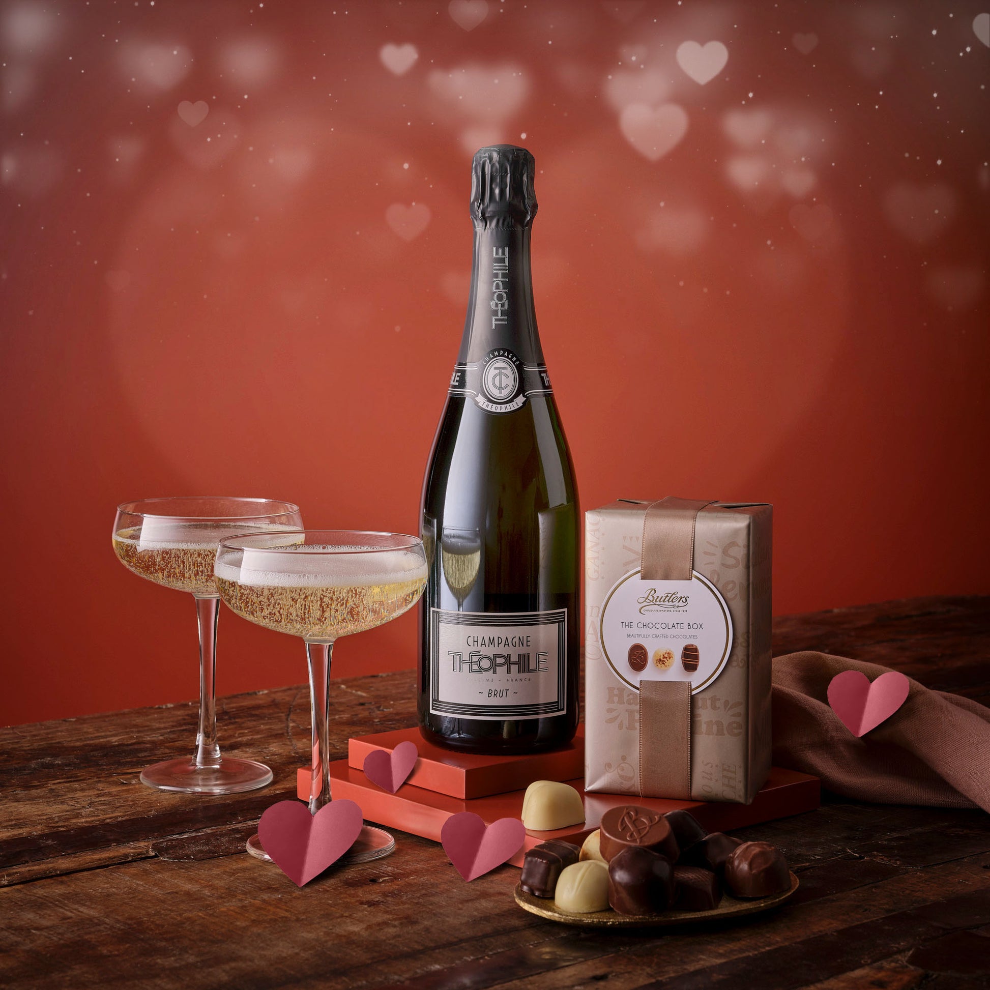 Champagne and chocolates for two hamper featuring a champagne bottle, two flutes, and a chocolate box on a wooden table with pink hearts.