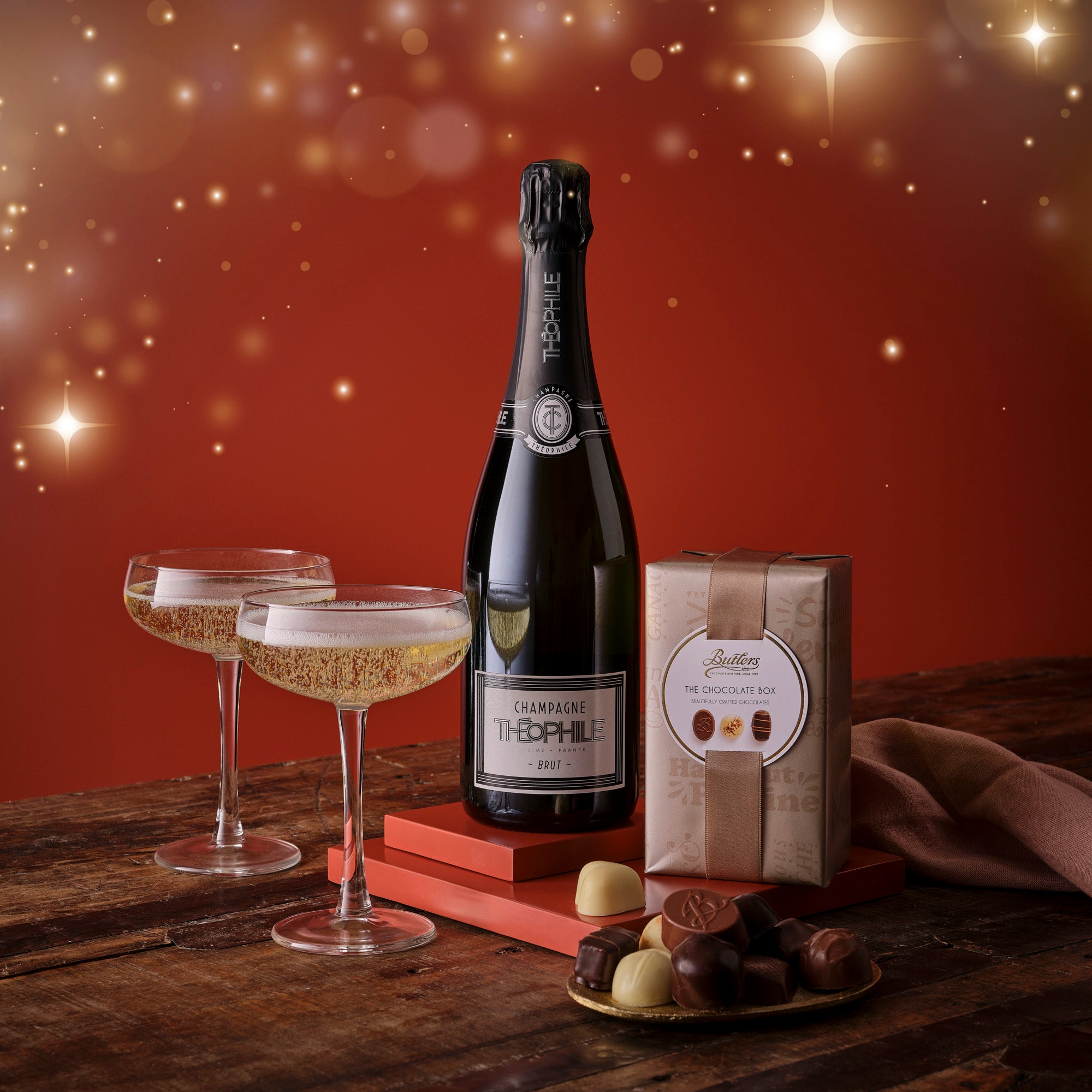 Bottle of champagne with two glasses and a box of chocolates on a wooden table against a warm, blurred background.