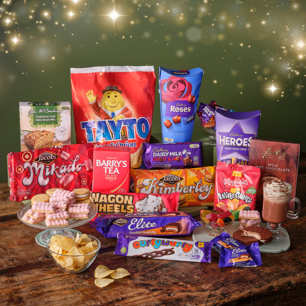 Assorted Irish snacks, Barry's Tea, Cadburys Roses, Tayto, Maynards jellies, wagon wheels, and Jacobs biscuits  on a wooden table with a festive background