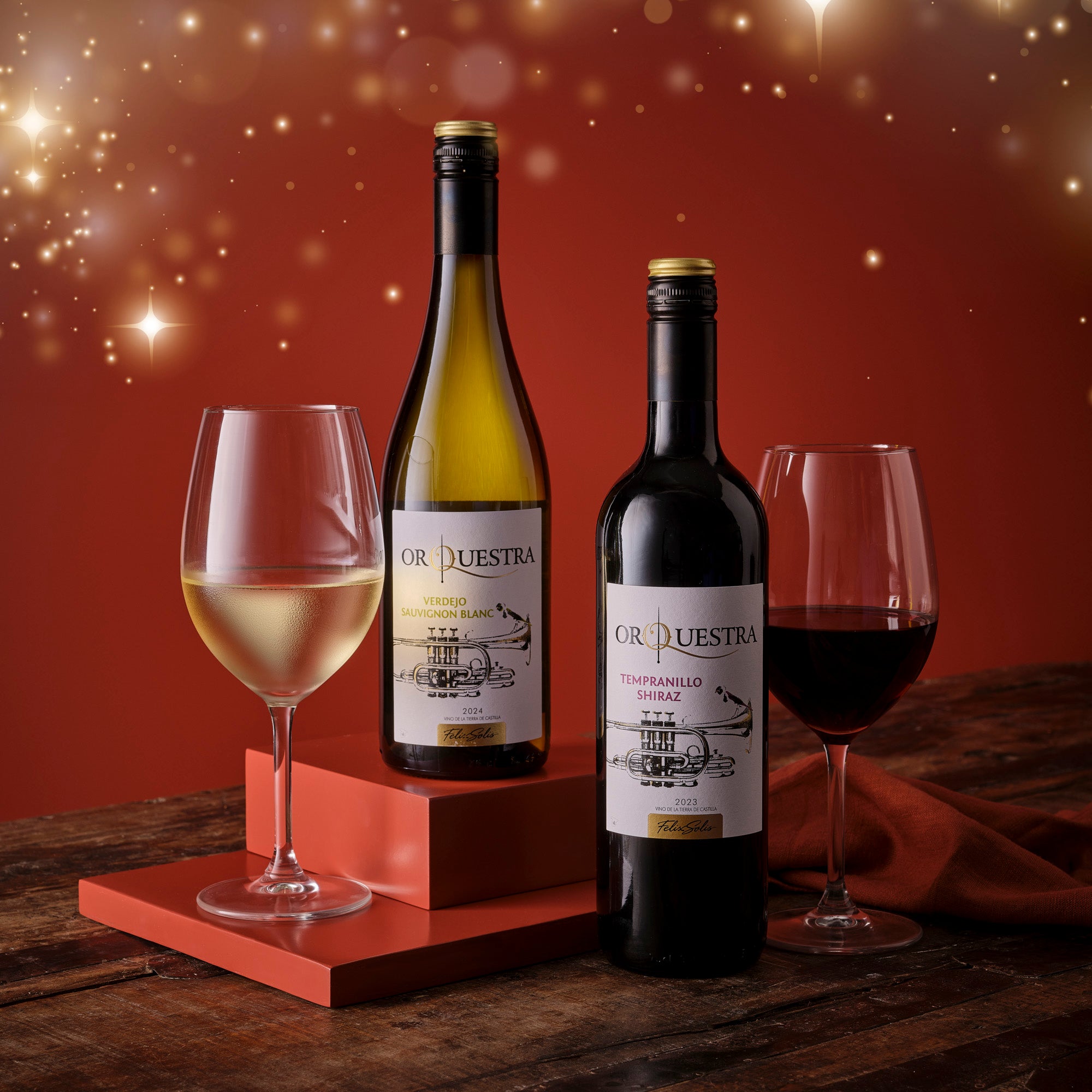 Two bottles of Orquestra wine with two glasses on a wooden surface against a red background.