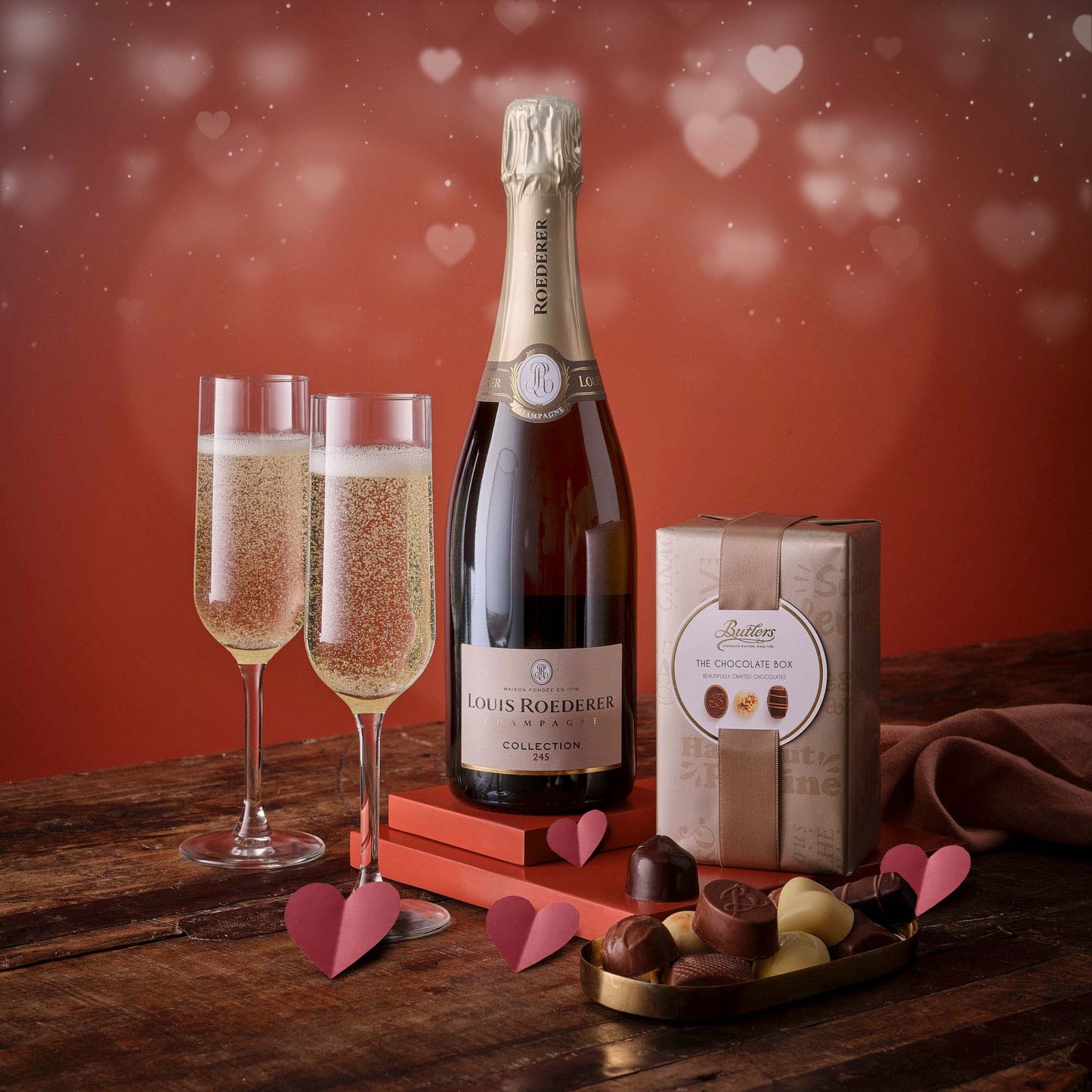 Luxury Valentine champagne and chocolate set with a Louis Roederer bottle, two flutes, a chocolate box, and assorted chocolates on a wooden table.