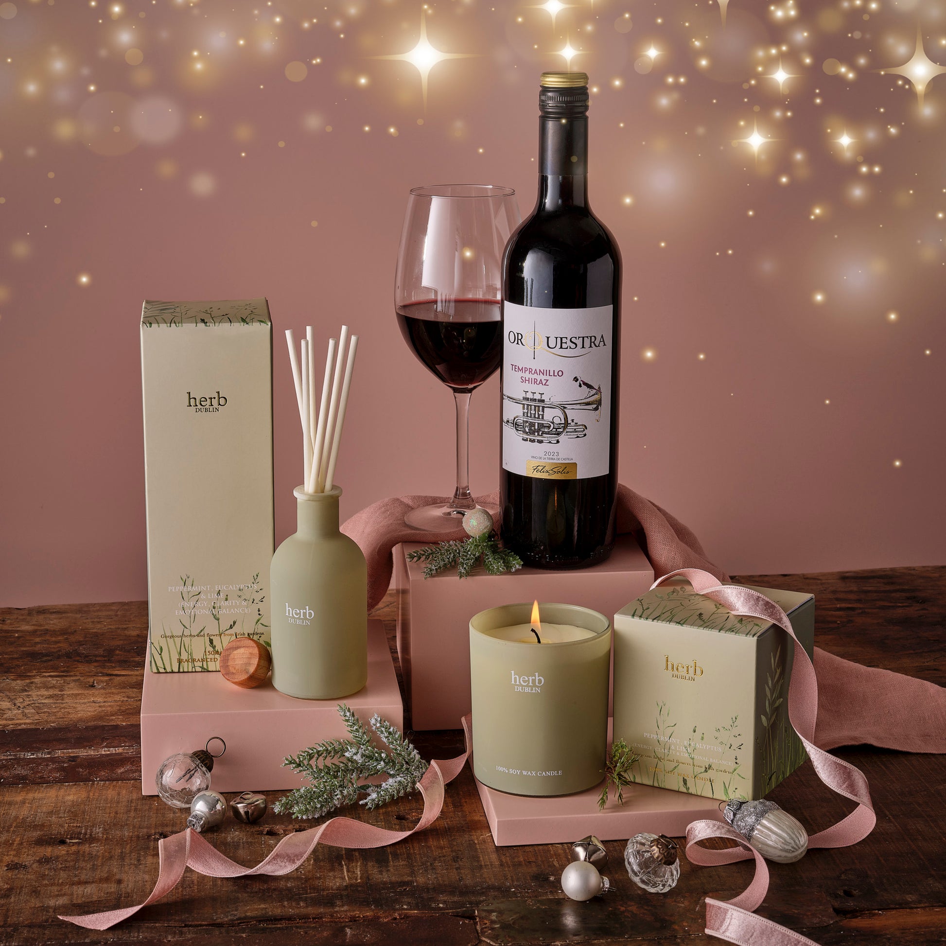 Gourmet gift set with  Herb Dublin diffuser, candle, bottle of red wine, and decorative items on a wooden table.