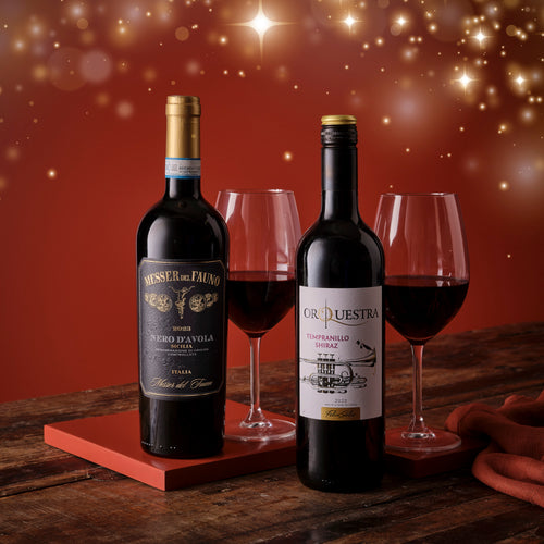 Two bottles of red wine with glasses on a wooden surface against a warm, blurred background.