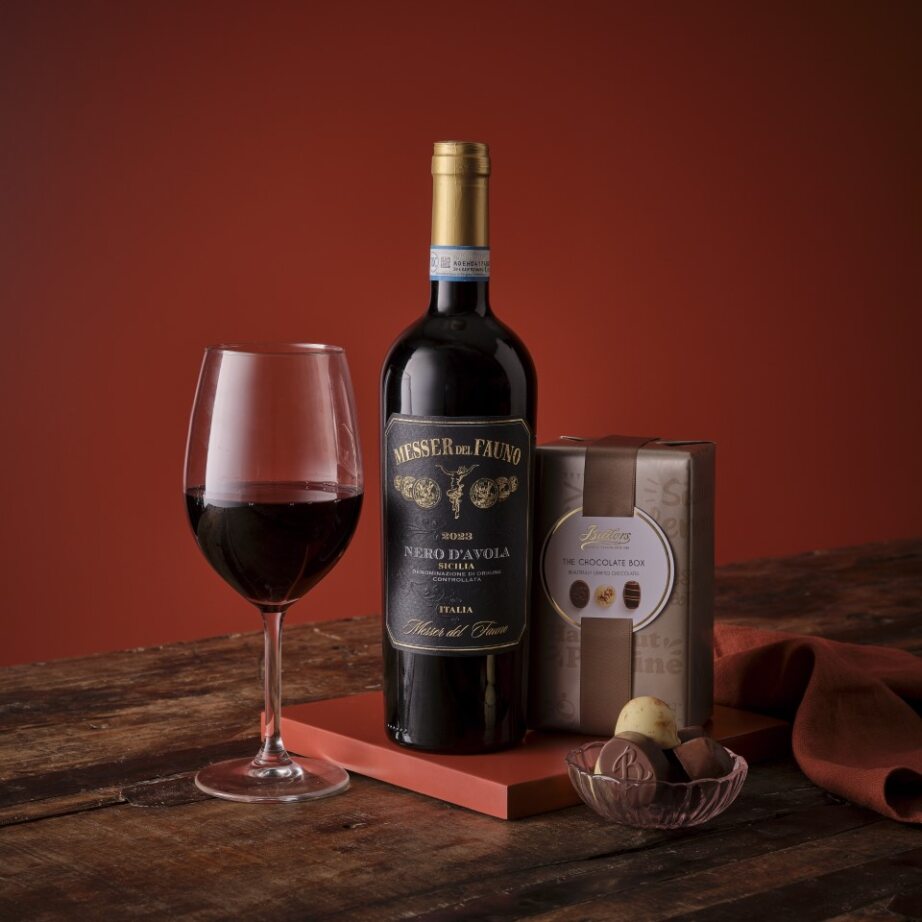 Bottle of red wine with a glass and box of chocolates on a wooden surface with a warm, blurred background.