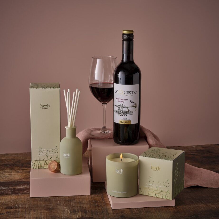 Relax and unwind hamper with red wine bottle, glass, diffuser and pastel gift boxes on a wooden surface.