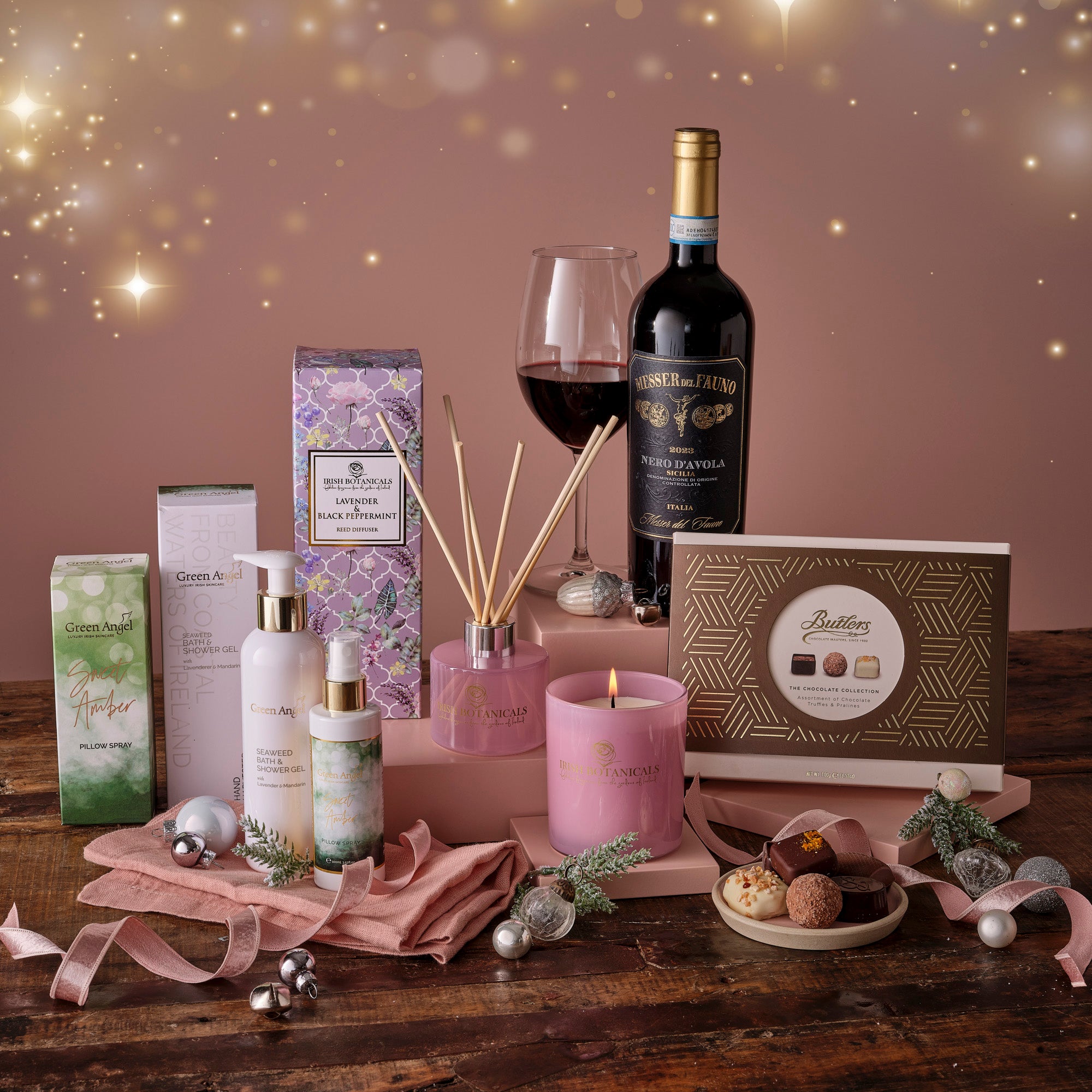 Gourmet gift set with green angel creams, diffuser, candle, wine, chocolate, and decorative items on a wooden table.