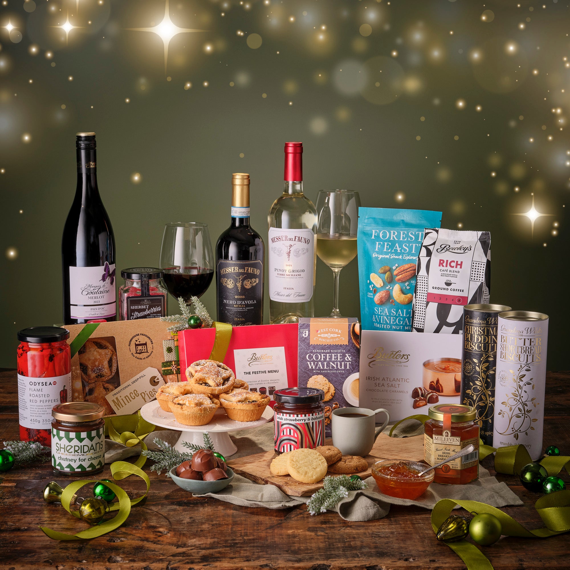 Gourmet gift set with red & white wines, snacks, and other treats on a wooden table with a festive background.