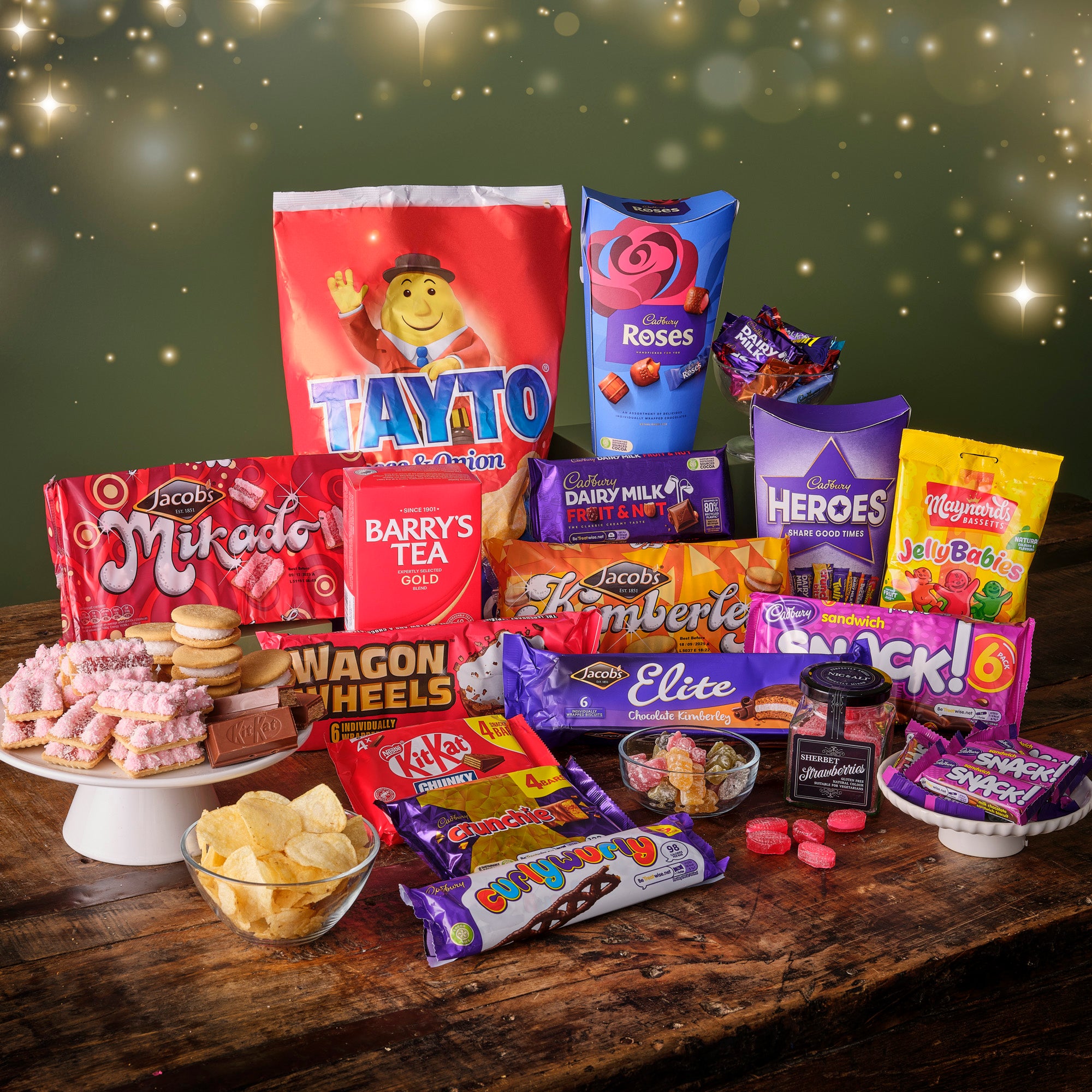 Assorted Irish snacks, Barry's Tea, Cadburys Roses, Tayto, Maynards jellies, wagon wheels, and Jacobs biscuits  on a wooden table with a festive background.