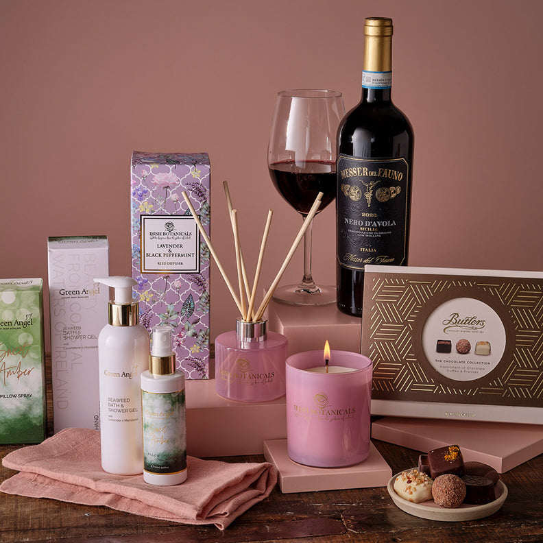Wine bottle, chocolates, candle and diffuser on a wooden table with decorative elements.
