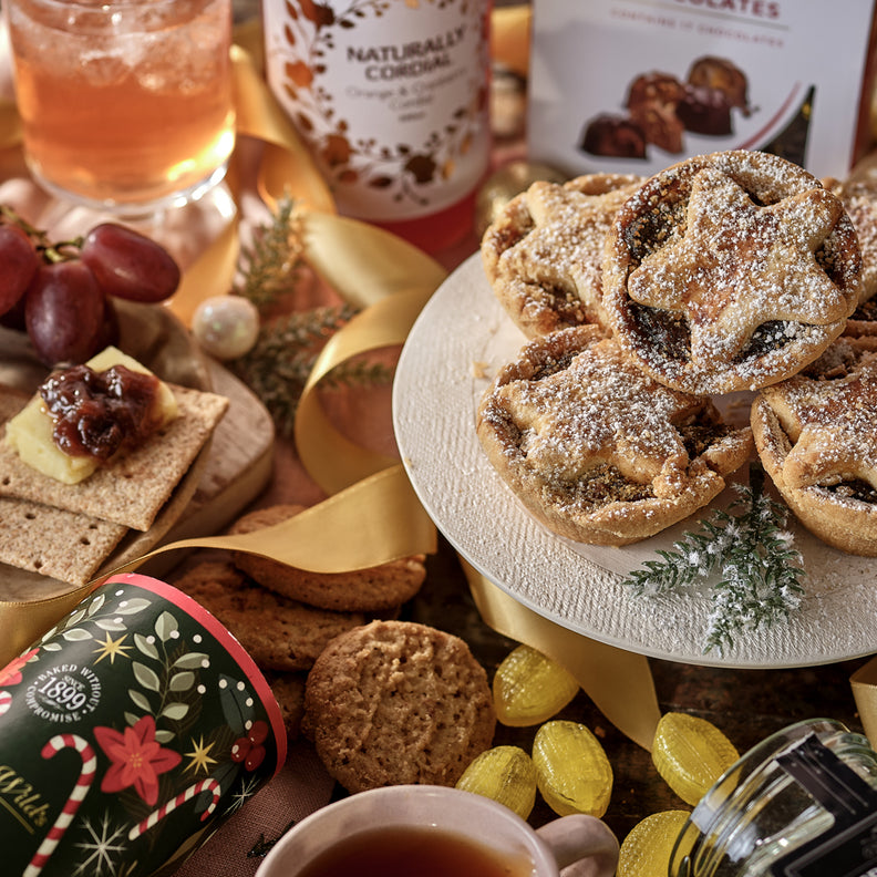 Assorted festive food and drink items including mince pies, cheese, and drinks on a wooden table.