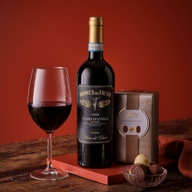 Bottle of red wine with a glass and chocolate box on a wooden surface with a warm, blurred background.