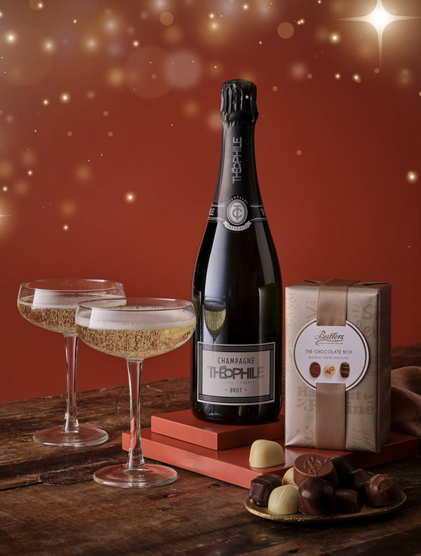 Bottle of champagne, two glasses, and a box of chocolates on a wooden table with a warm background.
