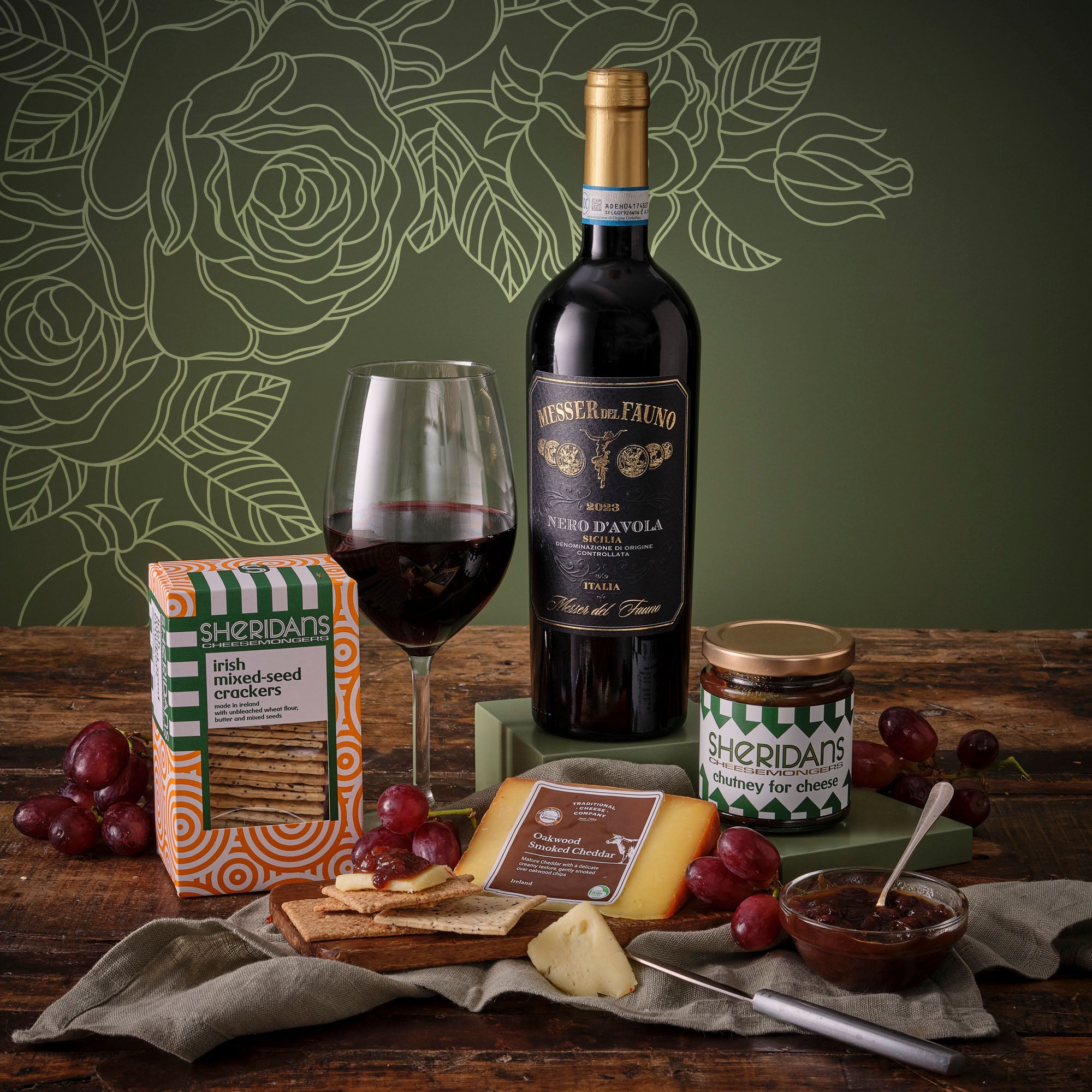 Mother’s Day Red Wine and Treats Hamper with a wine bottle, glass, crackers, cheese, chutney and grapes on a rustic table.
