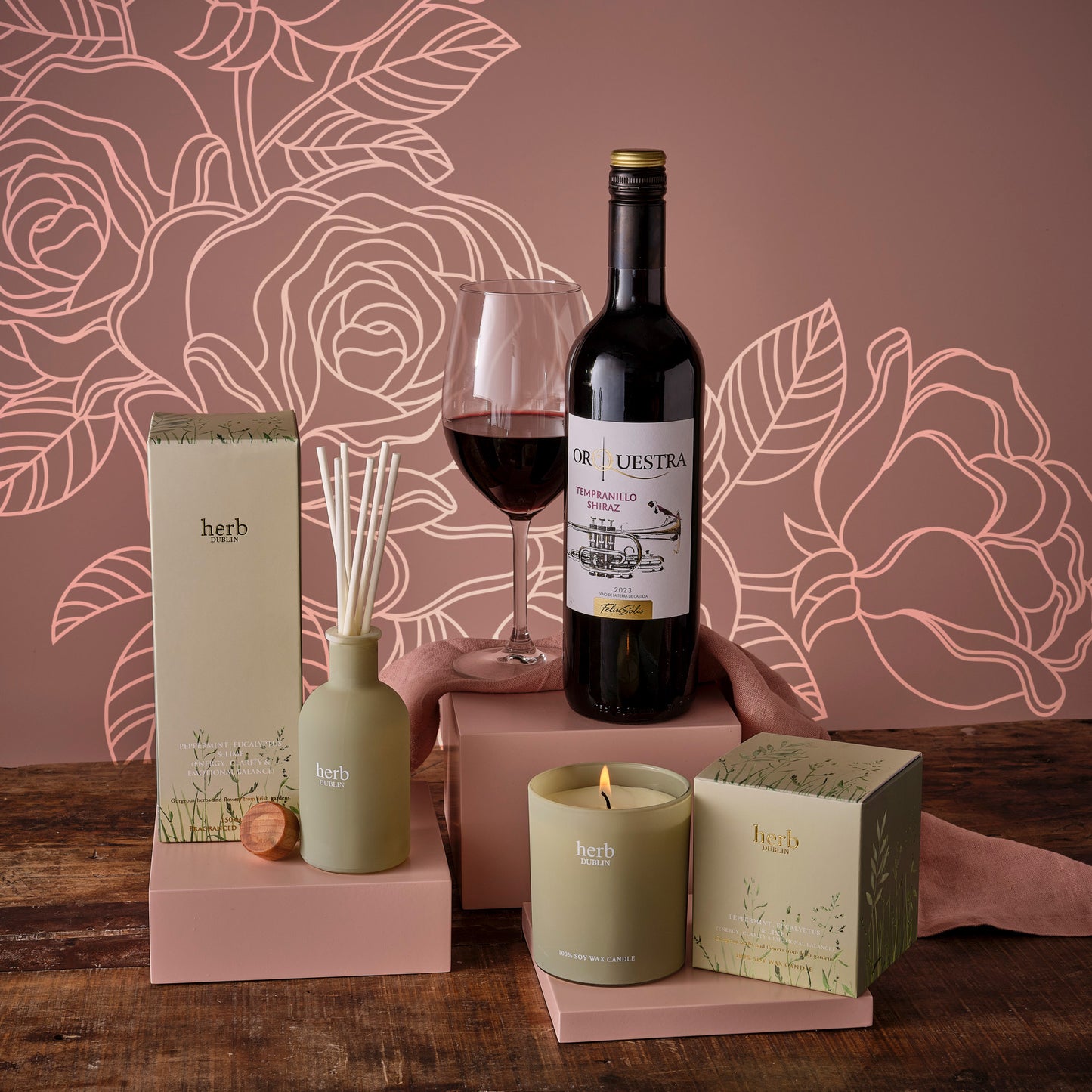 Mother's Day Relax Unwind Hamper includes red wine, a glass, diffuser, candles and treats on a pink display.
