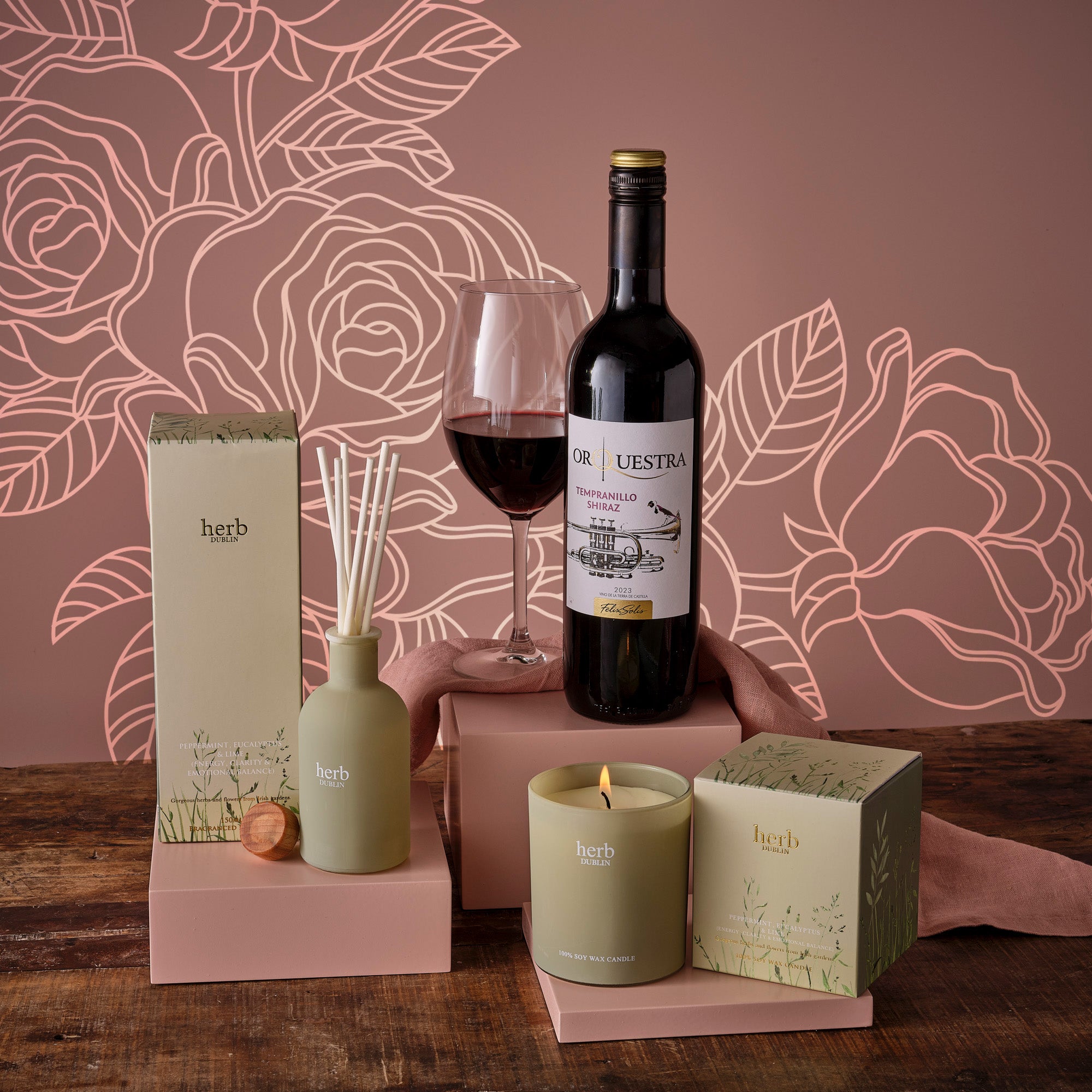 Mother's Day Relax Unwind Hamper includes red wine, a glass, diffuser, candles and treats on a pink display.