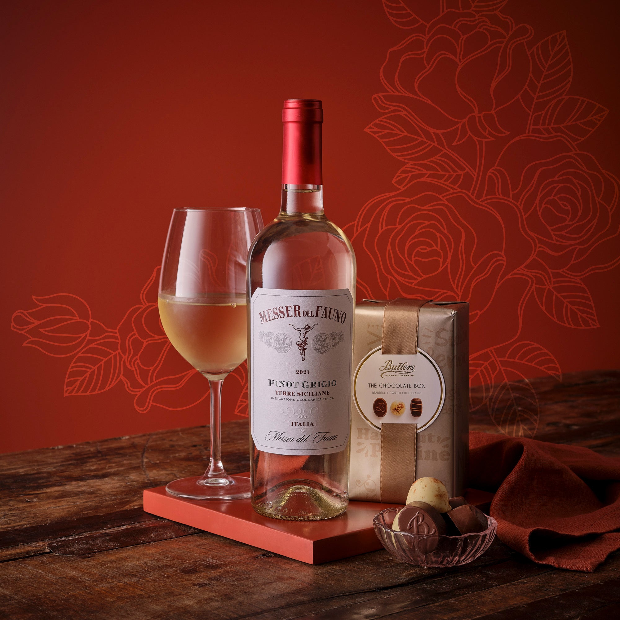 Pinot Grigio wine bottle with a glass of wine and a chocolate gift box on a wooden surface with a red floral backdrop